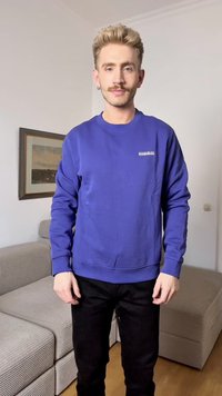Sweatshirt roxo com punhos e barras caneladas, apresentando um pequeno logotipo branco no peito. Usado com jeans pretos, ambientado em um ambiente interno.