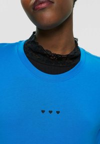Blå bomulds T-shirt med rund hals, med et sort hjertegrafik i midten. Blondekrave lagdelt under.