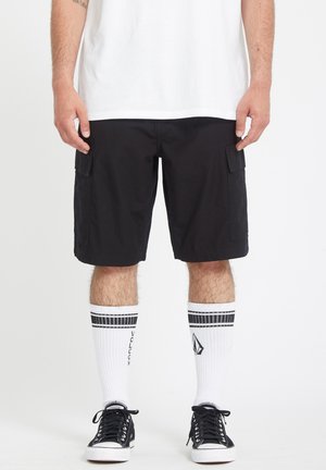 Homme portant un short cargo noir, des chaussettes blanches mi-mollet avec des bandes et logos noirs, ainsi que des baskets basses noires, debout avec les mains relâchées.