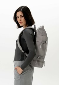 Zaino grigio con una forma aerodinamica e tessuto testurizzato, dotato di spallacci regolabili, fibbie laterali e chiusura con zip nella parte superiore.