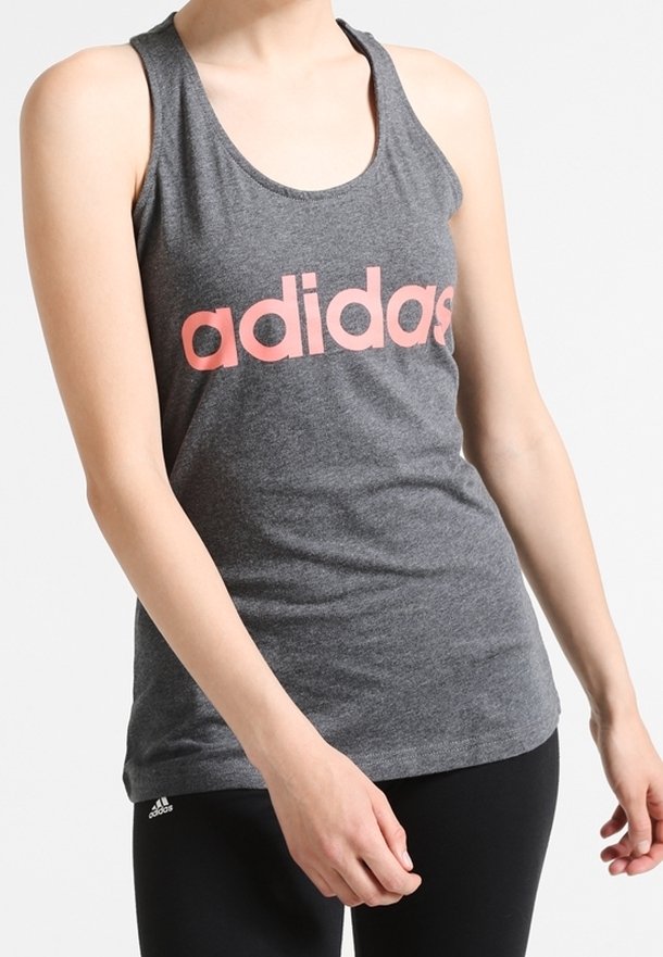 Persoon in een grijze Adidas tanktop met een roze logo en zwarte Adidas leggings tegen een effen lichte achtergrond.