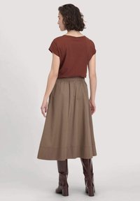 Nainen, jolla on lyhyet kiharat hiukset, päällä ruskea lyhythihainen paita, polven ylle ulottuva vaaleanbeige hame ja tummat ruskeat saappaat, seisoo selin.