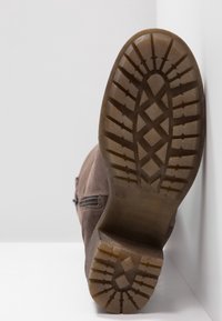 Semelle d'une botte en daim marron avec une semelle à motif et une fermeture éclair latérale, présentée à la verticale sur un fond blanc.