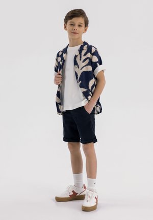 Kortärmad skjorta i marinblått med ett krämfärgat lövmönster, vit t-shirt, marinblå shorts och vita sneakers med röda detaljer, kombinerat med vita strumpor.