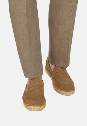 Onderbenen met lichtbruine broek en tan suède loafers met geweven jutezolen op een witte achtergrond.