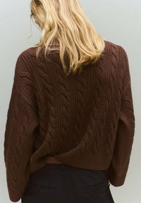 Brauner Strickpullover mit einem Zopfmuster, weiten Ärmeln und lockerer Passform. Die Textur wirkt weich und gemütlich, ideal zum Kombinieren.
