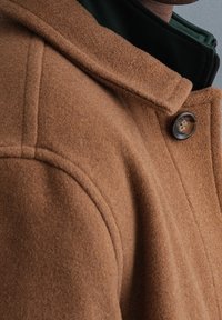 Brauner Wollmantel mit prominentem Kragen und einem dunklen Knopf. Die Textur wirkt weich und leicht flauschig, detailliert mit hellen Nähten.