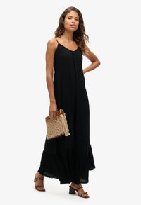 Zwarte maxijurk met een geplooide zoom, dunne bandjes en een vloeiende silhouet. Model houdt een geweven beige clutch vast en draagt tan blokhak sandalen.