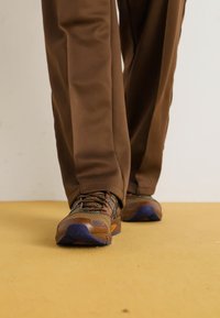 Rudi sportiniai batai su tinklelio panelėmis, tekstūruotais raišteliais ir violetiniu akcentu ant pado, derinami su rudomis kelnėmis stovinčioje pozicijoje.