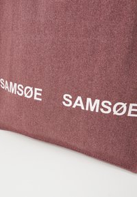 Gros plan d'un tissu rouge-brun texturé avec le texte blanc « SAMSØE » imprimé deux fois près du bord inférieur.