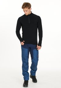 Zwart langemouw zip-up top gecombineerd met blauwe cargobroek met grote zakken en een klein logo aan de linkerkant. Zwarte laarzen.
