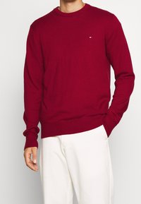 Maglione rosso lavorato a maglia con scollo rotondo classico, polsini e orlo a coste, caratterizzato da un piccolo logo sul petto. Abbinato a pantaloni bianchi.