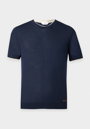 Hombre con camisa de manga corta azul oscuro, pantalones beige, gafas de sol oscuras y zapatillas bicolor, de pie con las manos en los bolsillos.