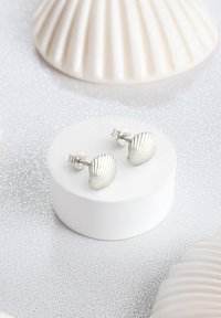 Silverfärgade snäckerformade örhängen med ribbad textur, med en klämbakdel. Visade på en rund vit pedestal, omgiven av mjuka, reflekterande ytor.