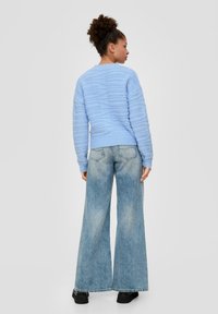 Lichtblauwe gebreide trui met textuurgolven, gecombineerd met wijde vervaagde blauwe jeans en zwarte enkellaarsjes, gezien van achteren.
