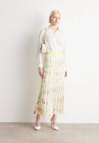 Blumige plissierte Midirock mit einem gelben Taillengürtel, kombiniert mit einer weißen Hemdbluse und einer cremefarbenen Handtasche. Die Absätze sind weiß.