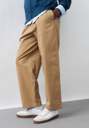 Osoba ubrana w beżowe spodnie o szerokich nogawkach, białe trampki z gumowymi podeszwami, szare skarpetki, białą koszulę i niebieską kurtkę, z rękami w kieszeniach.