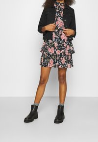 Svart denimjacka över en blommig tiered klänning med rosa och blå blommor på svart bakgrund, kombinerad med svarta ankelboots och grå strumpor.