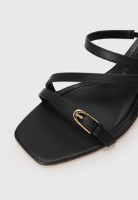 Black flat sandal with a square toe. Features crossed straps and a gold buckle accent. Made of smooth synthetic leather.