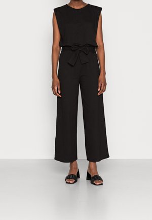 Schwarzer ärmelloser Jumpsuit mit tailliertem Gürtel und weit ausgestellter Silhouette; aus leichtem Stoff; kombiniert mit schwarzen Sandalen mit Absatz.