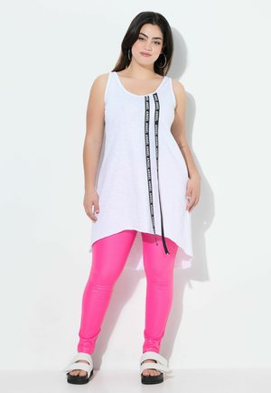 Túnica blanca sin mangas con un detalle de franjas laterales en negro, combinada con leggings rosa brillantes y sandalias blancas con una suela negra gruesa.