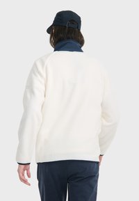 Weiße Fleecejacke mit strukturiertem Oberflächen, Reißverschlusskragen und marineblauen Akzenten. Kombiniert mit einer dunklen Kapuze, zeigt die Rückansicht Details der Ärmel und des Saums.