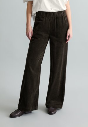 Person trägt dunkelbraune, weit geschnittene Cordhose, weißes eingestecktes T-Shirt und dunkelbraune Loafers vor einfachem Hintergrund.