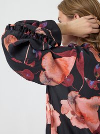 Czarna sukienka z przesadnie dużymi, falbaniastymi rękawami, zdobiona dużymi czerwonymi i różowymi kwiatowymi wzorami, wykonana z teksturowanej tkaniny, o luźnym kroju.