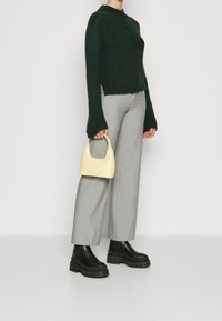 Pull en tricot vert, pantalon large gris, bottines chunky Chelsea noires et petit sac à main jaune avec une anse incurvée.