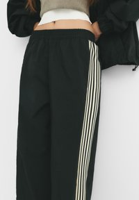 Pantaloni sport negri cu bandă elastică la talie, care au dungi verticale crem pe laturi, confecționați dintr-un material ușor și texturat.