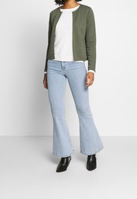 Green ribbed jacket over white textured top, blue flared jeans, and black ankle boots. Simple design with no visible fastenings or patterns.