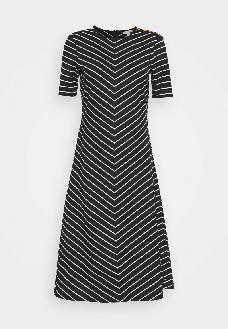 Robe noire trapèze avec des rayures diagonales blanches formant un motif en chevrons, manches courtes, et détails de boutons sur une épaule.