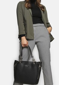 Vrouw in groene blazer en zwarte top, met een zwarte Guess-handtas, in combinatie met zwart-witte houndstooth broek.