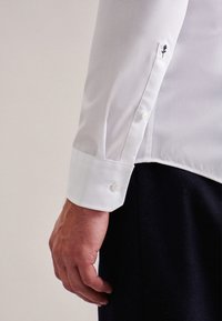 Wit overhemd van een gladde stof, met een knoopmanchet en een kleine zwarte rozenborduursel nabij de manchet.