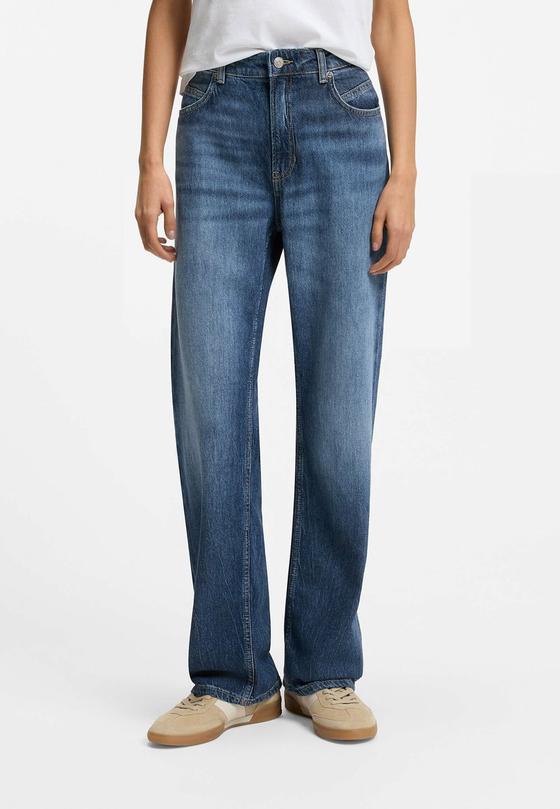 Personne portant un jean bleu taille haute à jambe droite, associé à des baskets beiges et une chemise blanche rentrée, debout sur un fond blanc.