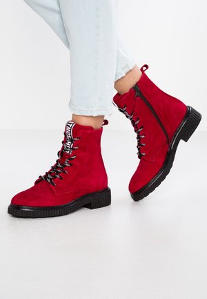 Punaiset mokkanahkaiset nilkkurit, joissa on nyöritykset, mustat pohjat ja sivuvetoketjut, pidetään vaaleansinisten lyhytlahkeisten farkkujen kanssa.