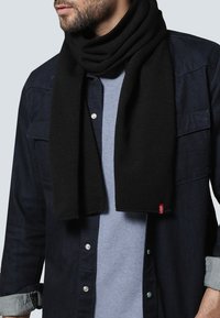 Bufanda de punto negra drapeada alrededor del cuello, con una textura suave y una forma rectangular limpia. Pequeña etiqueta de acento roja visible.