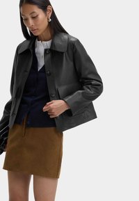 Kvinde iført en sort læderjakke over en navy cardigan og hvid skjorte, kombineret med en brun ruskinds mini-nederdel foran en ensfarvet baggrund.