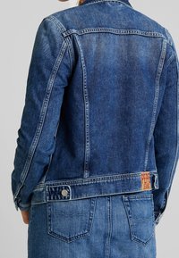 Denim jacket in dark blue with structured fit, visible stitching, and a branded tag on the back. Buttoned cuff details.