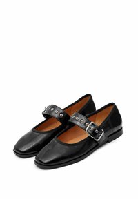 Ballerines en cuir verni noir avec un orteil carré, présentant une large sangle avec des œillets en métal et une boucle sur le côté. Texture lisse, design minimaliste.