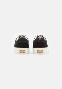Zapatillas de ante negras con costuras blancas y suela de goma beige. Presenta un logotipo en el talón. La vista trasera muestra la forma redondeada y detalles de diseño.