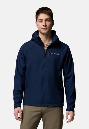 Junger Mann, der eine marineblaue Columbia-Zip-Jacke und beige Hosen trägt und vor einem schlichten hellen Hintergrund steht.