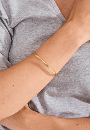 Ruka nosi tanku, uvijenu zlatnu narukvicu preko sive majice kratkih rukava, s rukom koja leži blizu prsa.