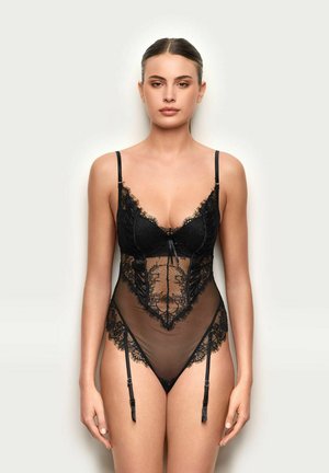 Mujer con cabello liso lleva un body de encaje negro transparente con tirantes de liga, de pie contra un fondo claro y liso, mirando de frente.