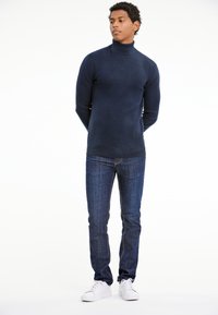Navyfarbener Rollkragenpullover in eng anliegender Passform, kombiniert mit dunkelblauen Jeans und weißen Sneakers. Glatte Textur in der gesamten Fläche.