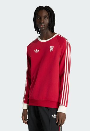 Persona de piel negra vistiendo un sudadera roja de Adidas con rayas blancas en las mangas y pantalones negros de Adidas, de pie frente a un fondo claro.