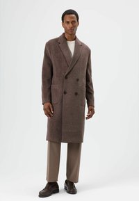Manteau marron en laine à double boutonnage avec deux poches avant, revers crantés et tissu texturé. Porté sur une chemise claire et un pantalon beige.