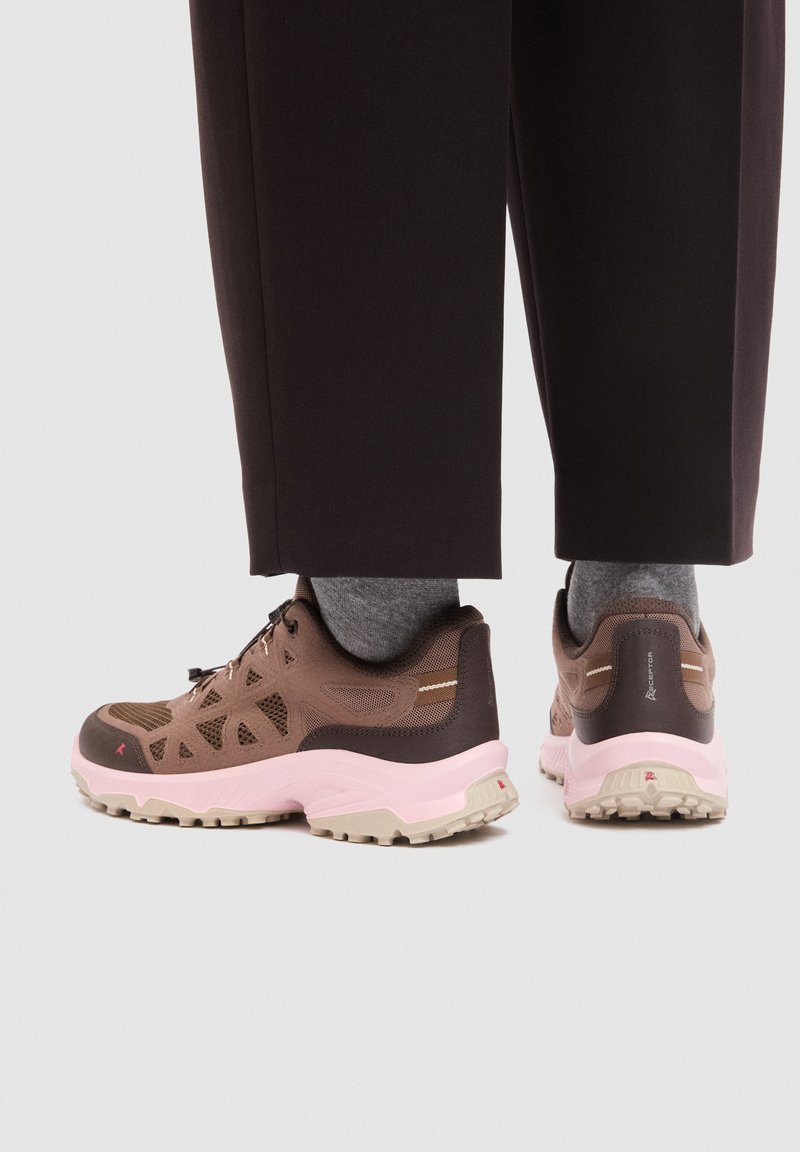 Baskets de trail marron et rose portées avec des chaussettes grises et un pantalon noir, vues de côté et de dos sur un fond clair.