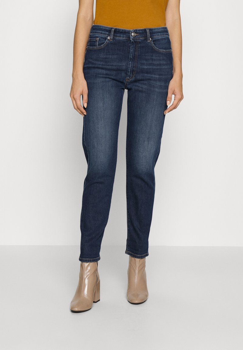 Tmavě modré džíny s rovnými nohavicemi, které mají jemné voskování, pět kapsový design a kontrastní švy, kombinované s béžovými kotníkovými botami.