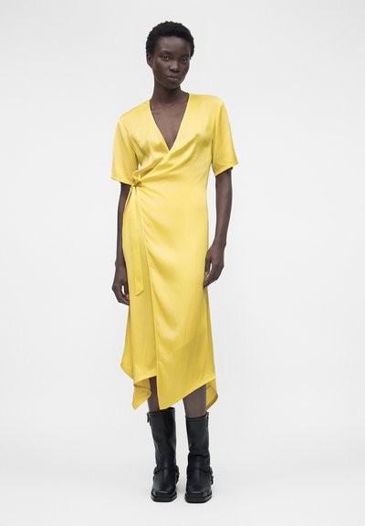 Γυναίκα στέκεται φορώντας κίτρινο φόρεμα τύπου wrap με κοντά μανίκια και μαύρες μπότες μέχρι τη μέση της γάμπας σε λευκό φόντο.